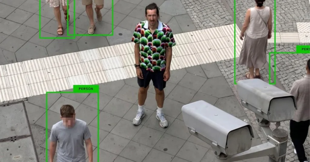 Surveillance-style object-detection overlay marking every pedestrian with a green PERSON bounding box except the wearer in Simon Weckert's Digital Camouflage patterned shirt
